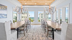 A dining area with a long wooden table, white cushioned chairs, chandeliers, and large windows offering ocean views. - Emerald View|Easy Beach Access|Unobstructed Gulf Views|Private Pool (Inlet Beach)