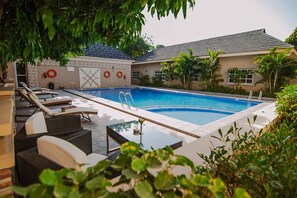 Piscina con sillas de descanso y una mesa, rodeada de follaje y un edificio con techo de tejas.