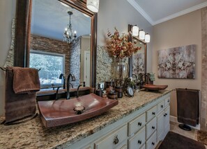 Une salle de bain avec un grand miroir, un lavabo marron, un lustre, un vase de fleurs séchées et un tableau au mur.
