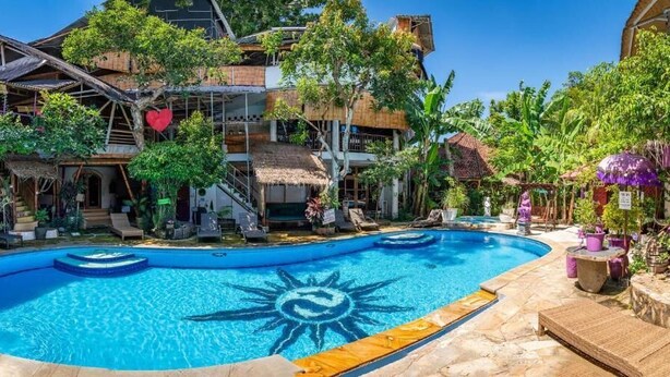 A swimming pool with a sun design, surrounded by tropical plants and a multi-story building with a thatched roof. - Seabreeze Hostel Bali (Canggu)