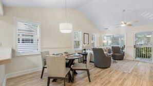 Amplia sala de estar con comedor, ventanas grandes con persianas, ventilador de techo y vistas al exterior.