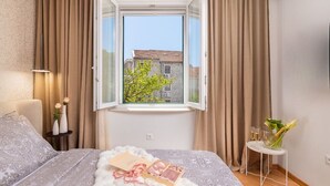 A bedroom with a large bed, a window with curtains, a bedside table with a lamp, and a small table with a vase and glasses. - Luxury Apartment Luft Split Near Old Town (Split)