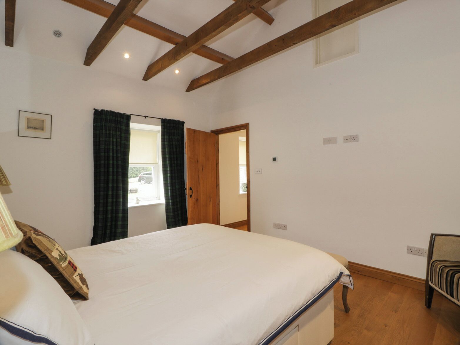 A bedroom with a large bed, wooden beams on the ceiling, a window with curtains, and a door leading to another room.