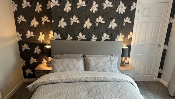 A neatly made bed with a grey headboard, two bedside tables with lamps, and a dark wall with a leaf pattern. - Cosy Cottage Apartment (Argyll and Bute Council)