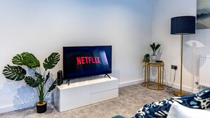 A modern living room with a flat-screen TV displaying 'NETFLIX' in red letters, a white entertainment unit, a large potted plant, a floor lamp, and a patterned sofa. - Spacious & Modern 2-Bedroom House (Worksop)