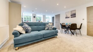 A modern living room with a blue sofa, a dining table with chairs, and a kitchen in the background. - Spacious & Modern 2-Bedroom House (Worksop)