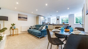 A modern living room with a blue sofa, dining table, and chairs. A kitchen is visible in the background with a window providing natural light. - Spacious & Modern 2-Bedroom House (Worksop)