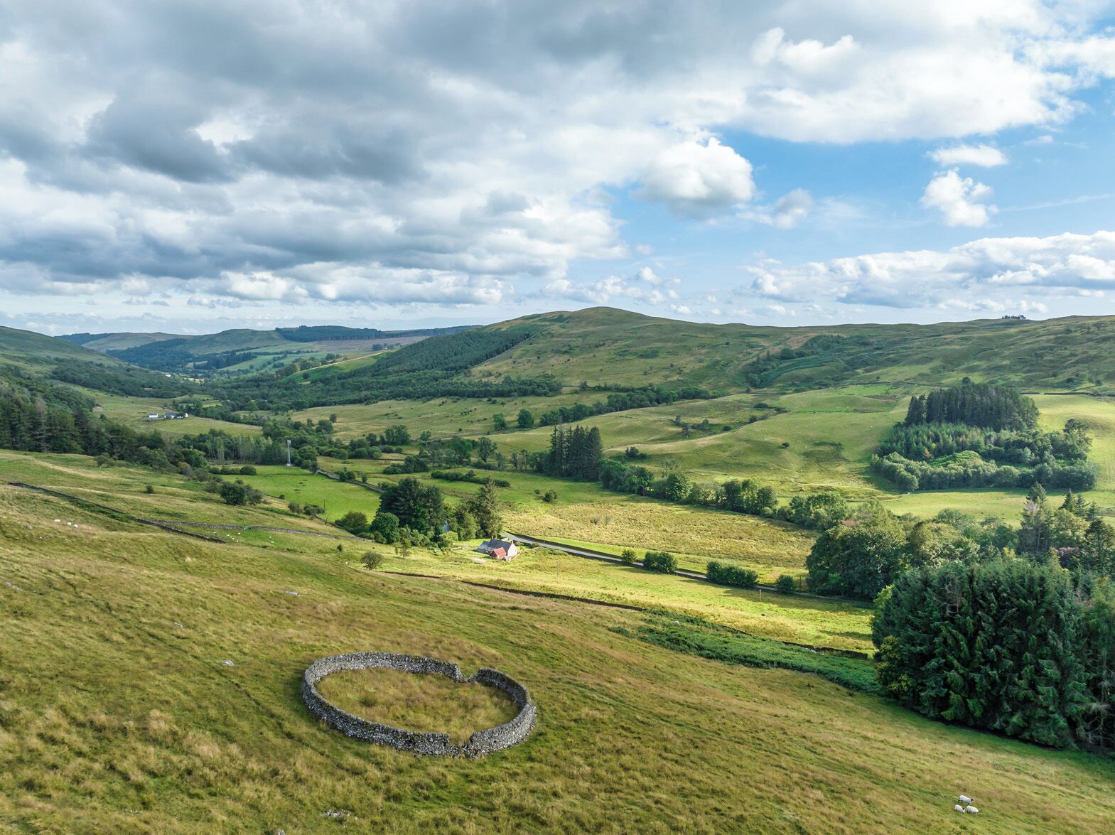 Paisaje rural con colinas onduladas, un círculo de piedras y una casa pequeña a lo lejos.