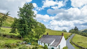 A white cottage with a stone roof, surrounded by a stone wall and a wooden fence, set in a lush green landscape with a winding road. - High Auchenbrack (Thornhill)