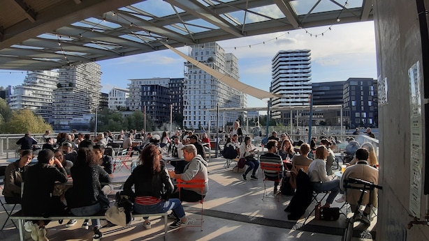 Un espacio al aire libre con mesas y personas cenando, con vistas a un paisaje urbano de edificios modernos.