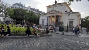 Una calle adoquinada con gente sentada y de pie alrededor de un edificio con columnas y banderas.