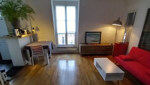 Una sala de estar con piso de madera, un sofá rojo, una mesa de centro blanca, una zona de comedor con mesa y sillas, un televisor sobre un mueble bajo, y un ventanal con vistas.