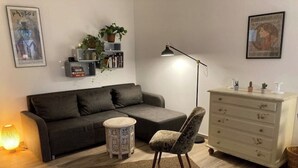 A living room with a grey sofa, a patterned armchair, a small round table, a floor lamp, and a white dresser with decorative items.