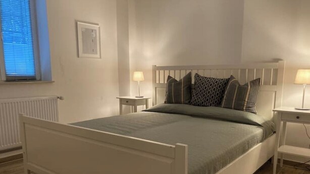 A bedroom with a white bed, two bedside tables with lamps, a window with blinds, and a radiator.