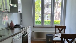 A modern kitchen with white appliances, a window with a view of greenery and buildings, and a dining area with a wooden table and chairs. - Living in the Mayor‘s Mansion (Hamburg)
