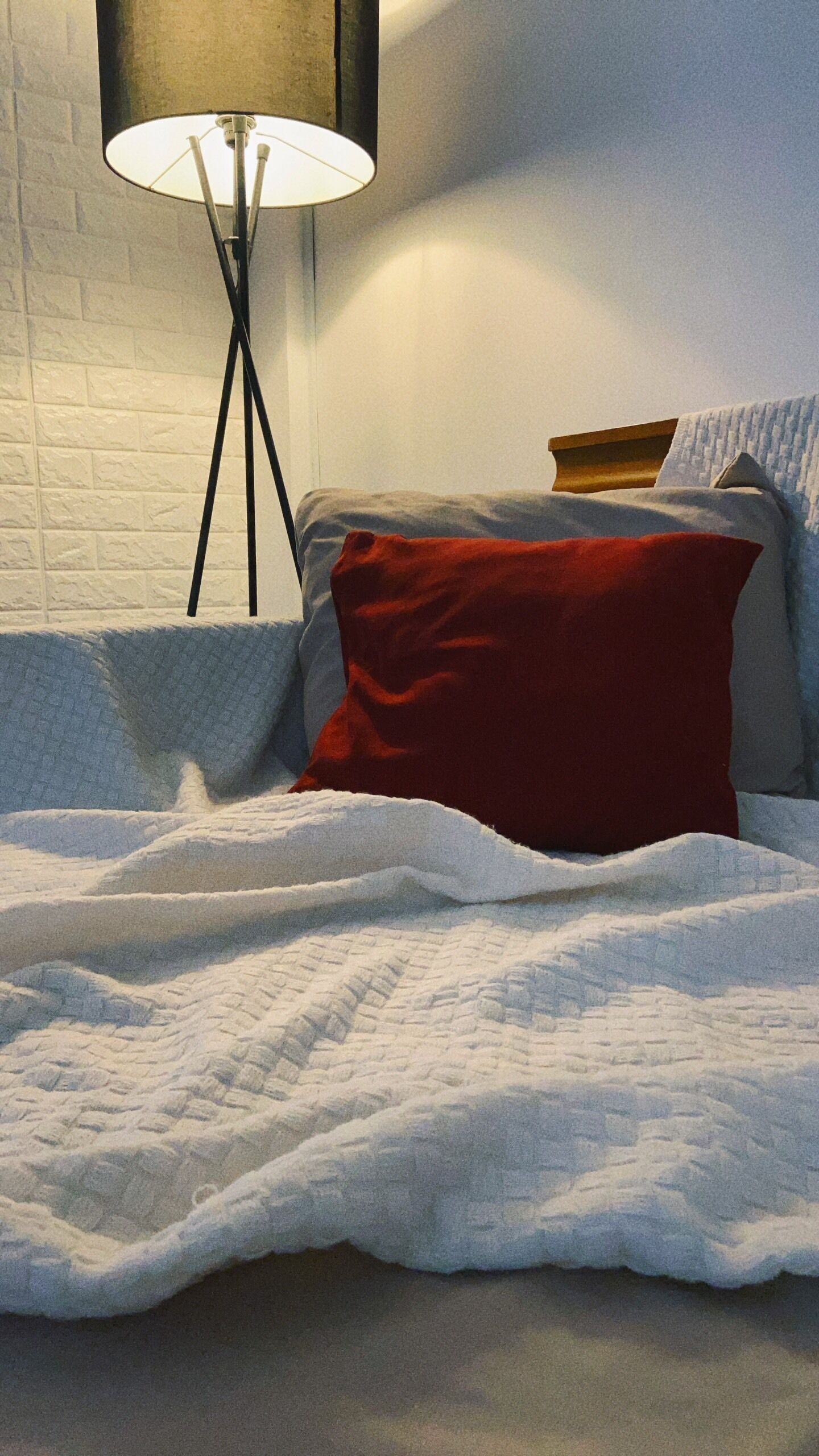 A neatly made bed with a white quilt, a red pillow, and a floor lamp with a white shade.