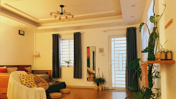 A spacious living room with a sofa, a coffee table, and a dining area. There are two windows with curtains, a plant, and a painting on the wall. - Memories about Dalat in Indochine (Da Lat City)