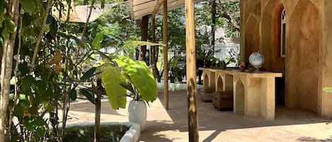 A covered outdoor area with a concrete floor, surrounded by greenery and a building with arched windows.