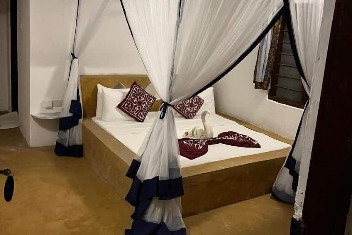 Una habitación con cama bajo un dosel, una mesita con una silla y una ventana con cortinas.