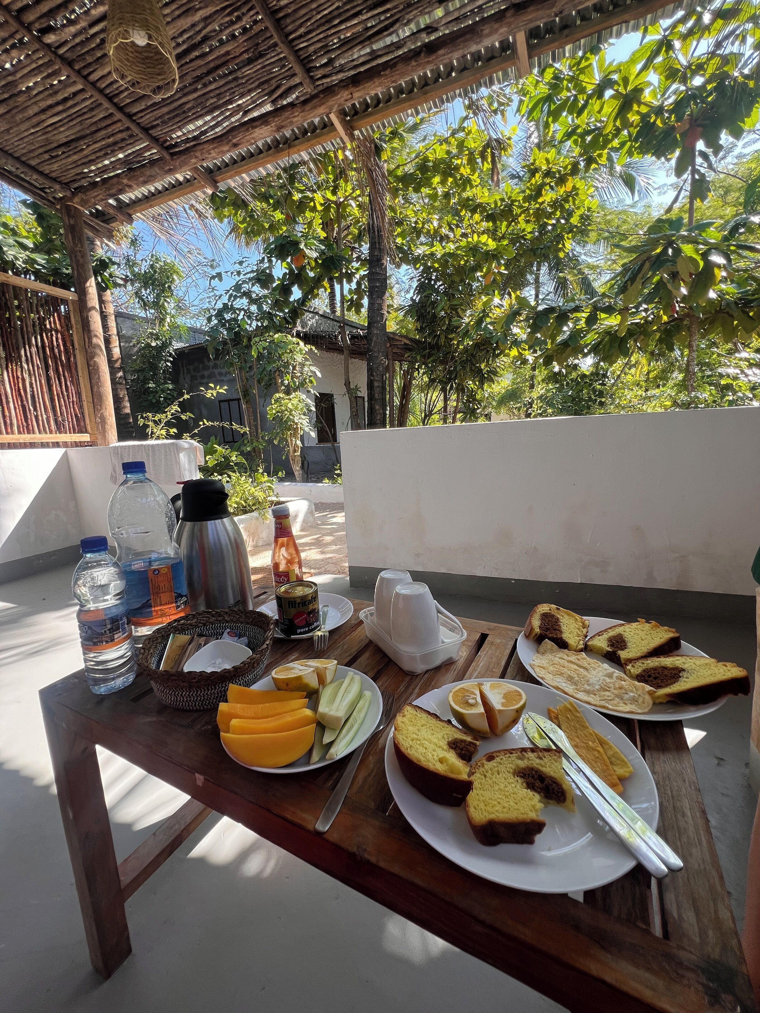 Una mesa de madera con alimentos para el desayuno, como pan, fruta y bebidas, ubicada al aire libre con vistas a árboles y un edificio al fondo.