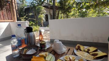 Una mesa de madera con alimentos para el desayuno, como pan, fruta y bebidas, ubicada al aire libre con vistas a árboles y un edificio al fondo.
