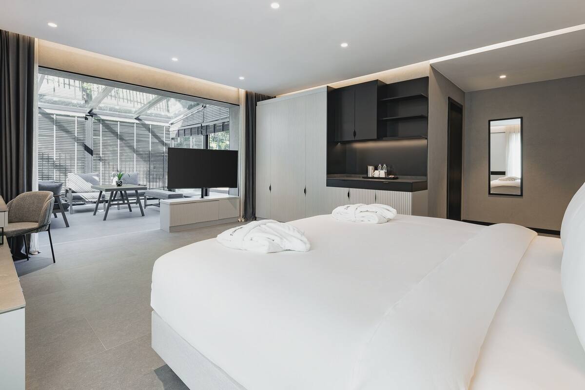 Ein modernes Hotelzimmer mit einem großen Bett, einem Schreibtisch mit Stuhl, einem Fernseher und Blick ins Freie durch große Fenster.