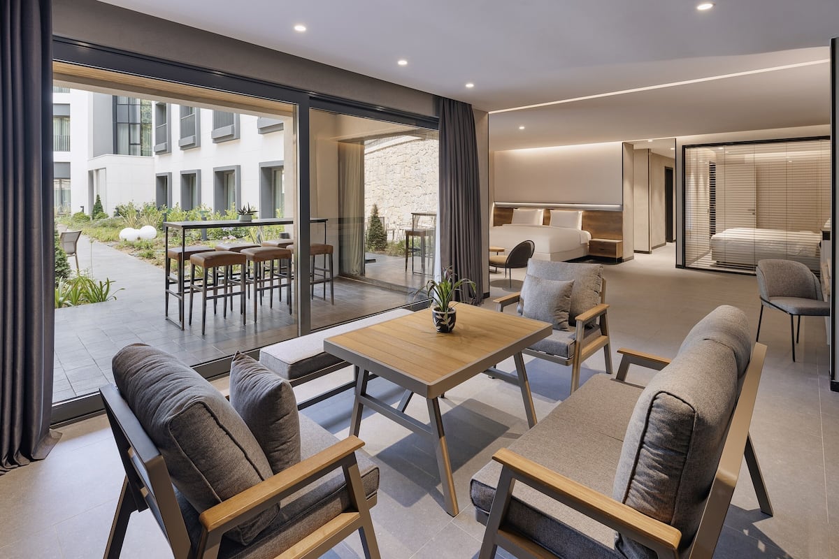 Ein modernes Hotelzimmer mit einer großen Schiebeglastür, die auf eine Terrasse im Freien führt, einer gemütlichen Sitzecke mit Sofas und einem Holztisch sowie einem separaten Schlafbereich mit Bett und Fenster.