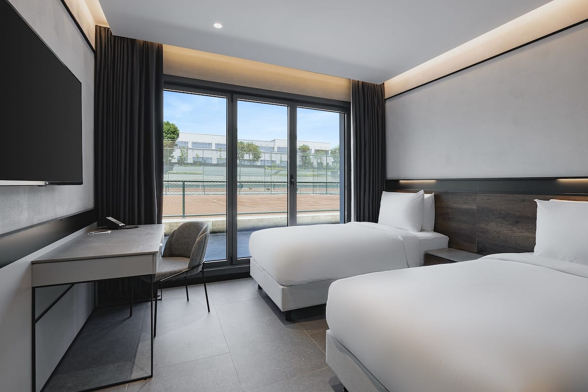 Ein modernes Hotelzimmer mit großem Fenster, Schreibtisch, zwei Betten und einem Sessel.