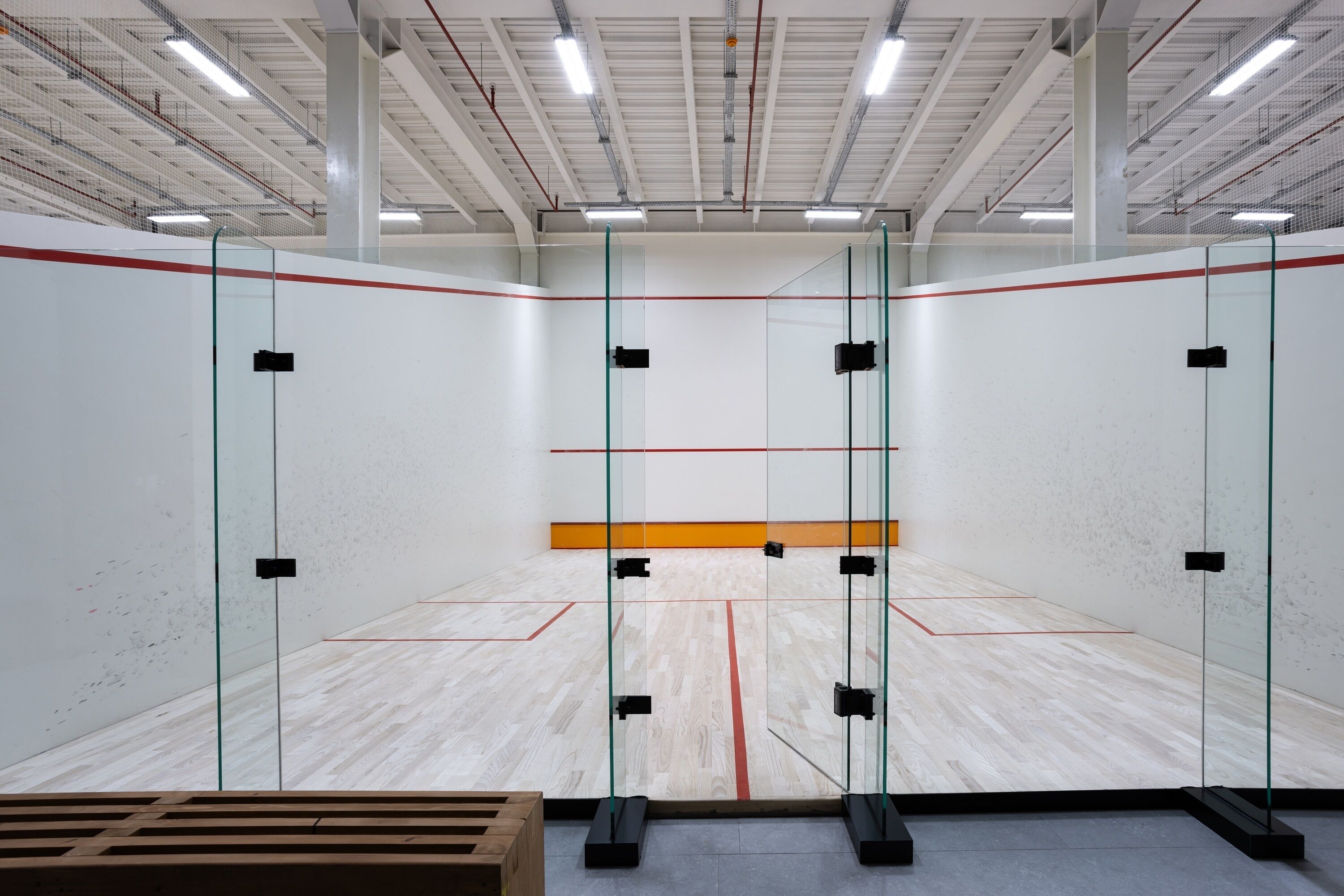 A squash court with glass walls, a wooden floor, and a red line marking the playing area.