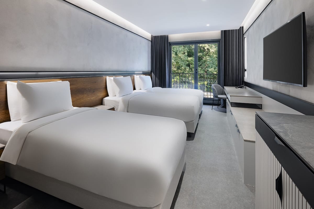 Ein modernes Hotelzimmer mit zwei Betten, einem Schreibtisch, einem Flachbildfernseher und einem Balkon mit Blick auf Bäume.