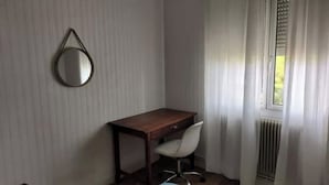 Une petite pièce avec un bureau en bois, une chaise blanche, un miroir rond accroché au mur et une fenêtre avec des rideaux.