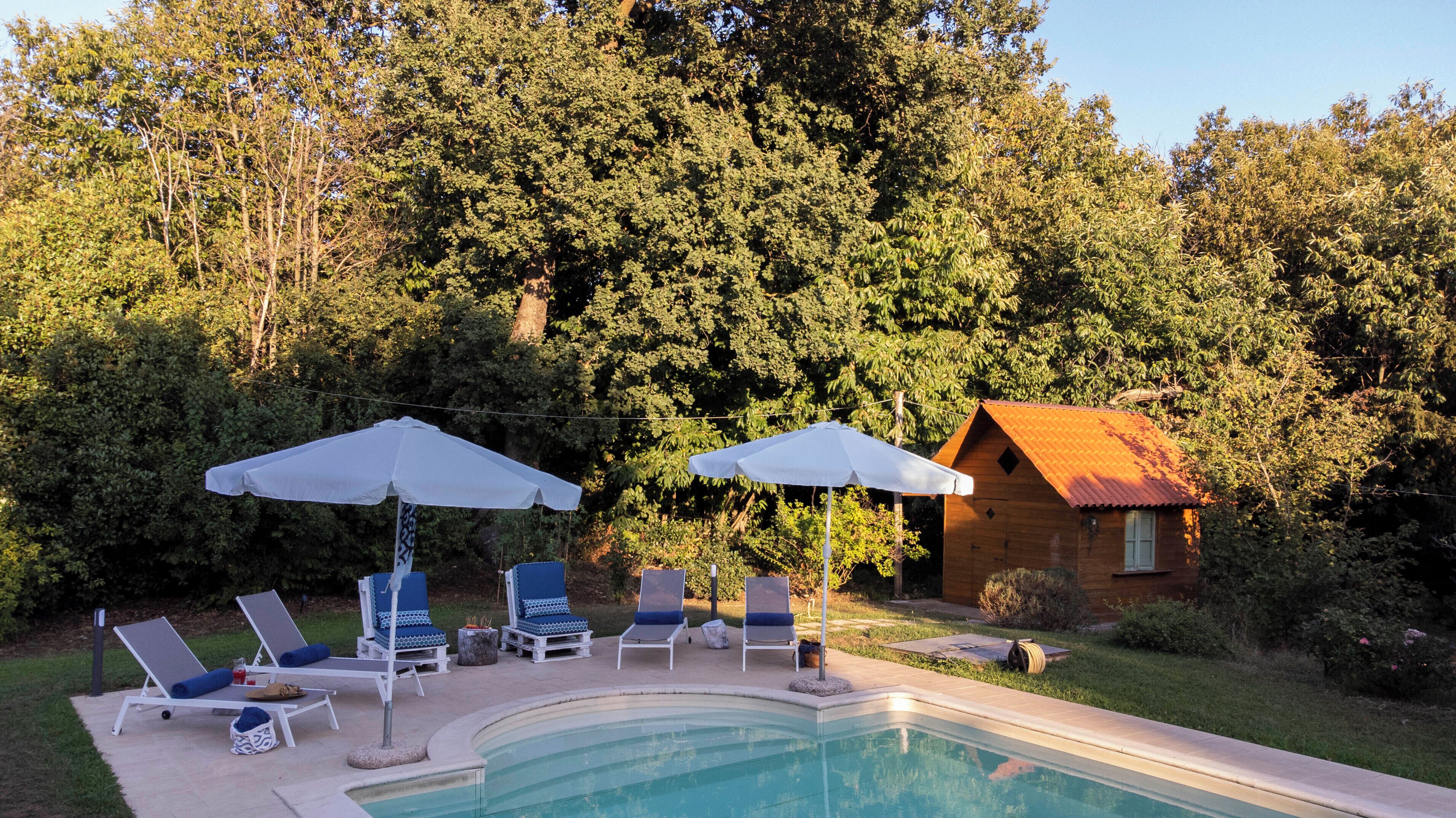 Area piscina con lettini e ombrelloni, circondata da alberi e un rifugio in legno.