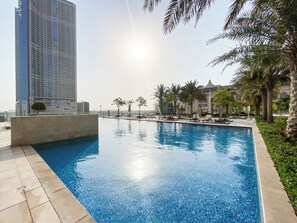 A swimming pool with lounge chairs and palm trees, surrounded by a building with a dome and a tall skyscraper in the background.