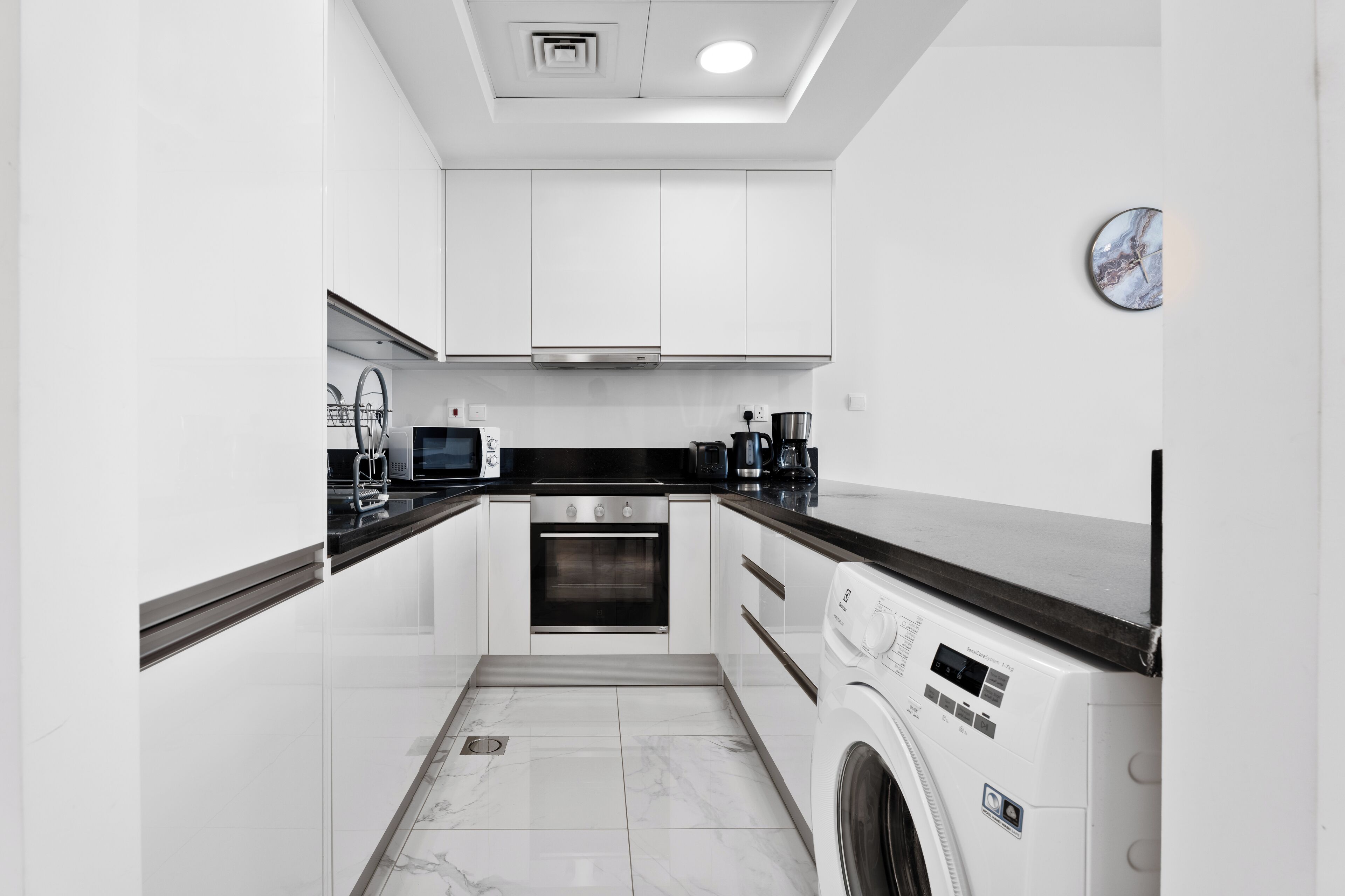 A modern kitchen with white cabinets, a built-in oven, a microwave, and a washing machine.