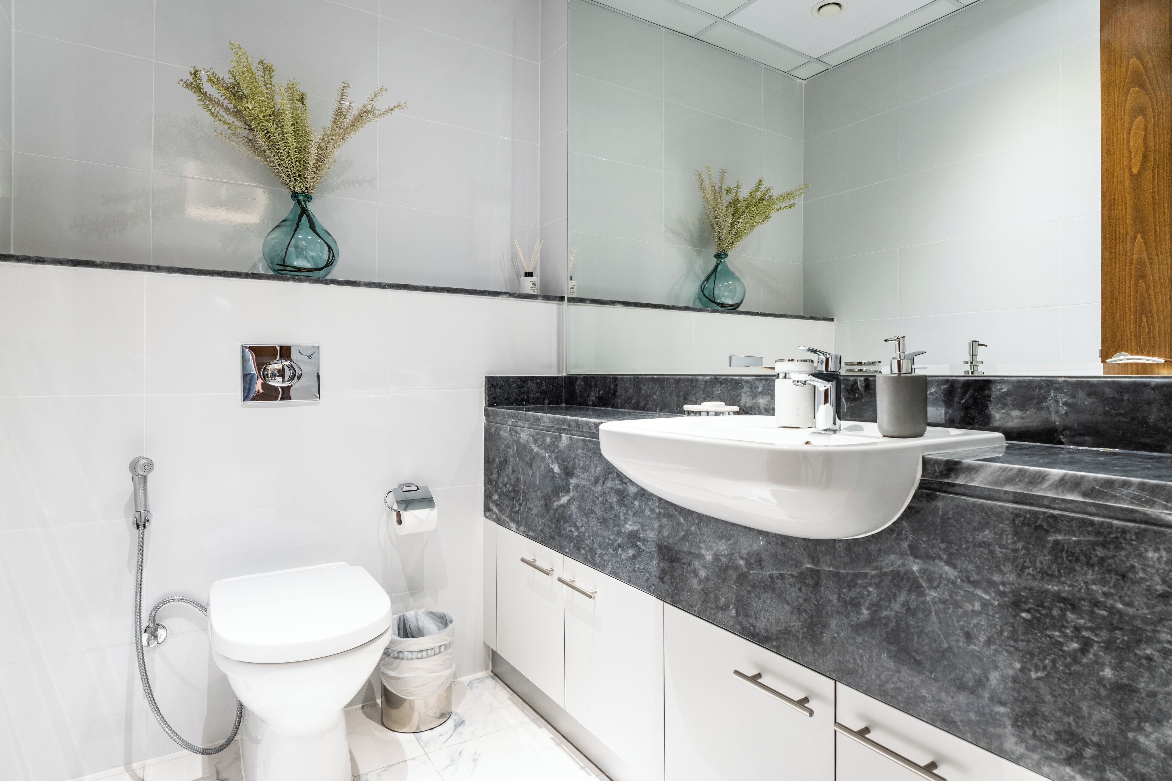 Un baño moderno con inodoro blanco, lavamanos con encimera de mármol y dos jarrones con plantas secas en la pared.