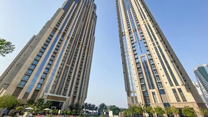 Due edifici residenziali alti, con diversi piani, una zona verde alberata e una piscina, con cielo sereno.