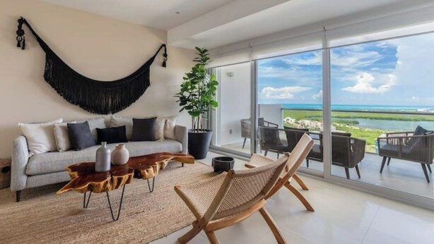 Ein modernes Wohnzimmer mit einer Couch, einem Couchtisch und Korbstühlen, mit Blick auf eine malerische Meereslandschaft.