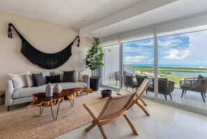 Relax in this stylish living room with a sofa, coffee table, and woven chair, opening to a balcony with a view of lush greenery.