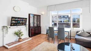 A modern living room with a flat-screen TV, a dark wooden cabinet, a dining area with chairs, a sofa, and a large window offering a view of buildings. - Duplex 3 Bedroom Flat Golders Green Central London (London)