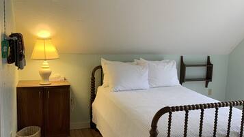 Un dormitorio con una cama de madera, una mesita de noche con lámpara, un armario de madera y un pequeño cubo de basura.