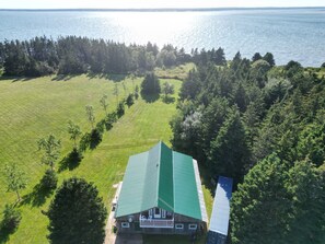 A large house with a green roof, surrounded by a grassy field and dense forest, overlooking a body of water. - The Barn at Spry Point (Little Pond)