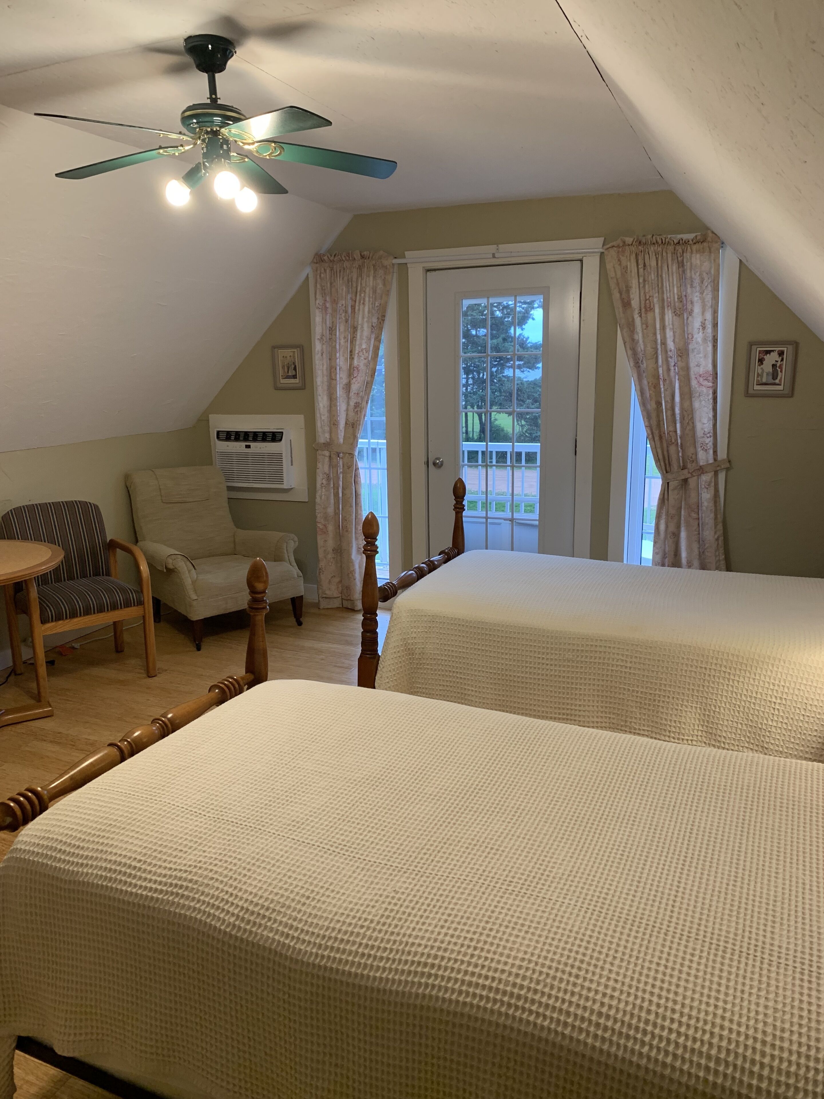 Una camera da letto con due letti, un ventilatore a soffitto, una sedia, un condizionatore d'aria e una porta che dà su un balcone.