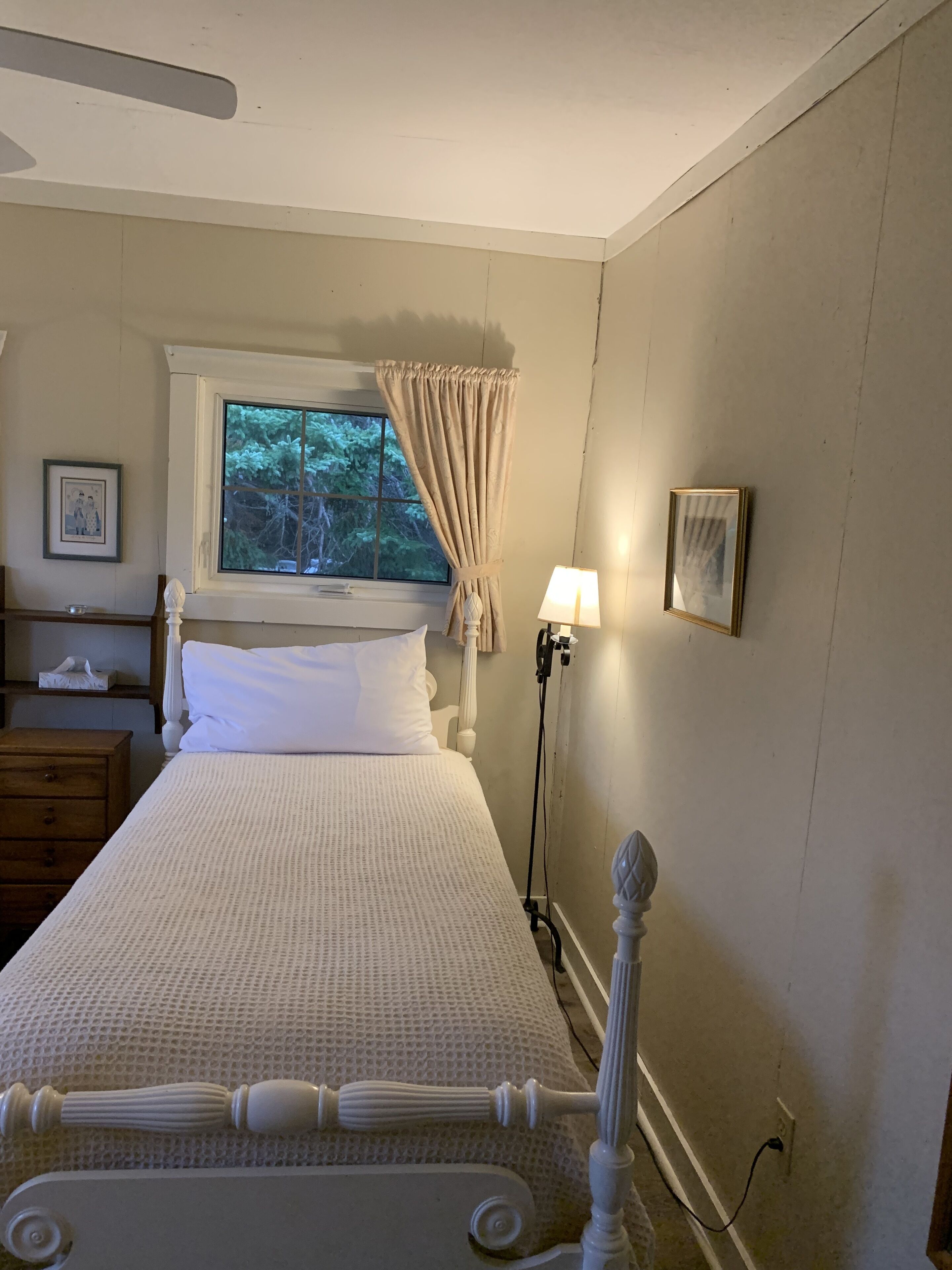 A bedroom with a bed, a bedside table, a lamp, a window with curtains, and a picture on the wall.