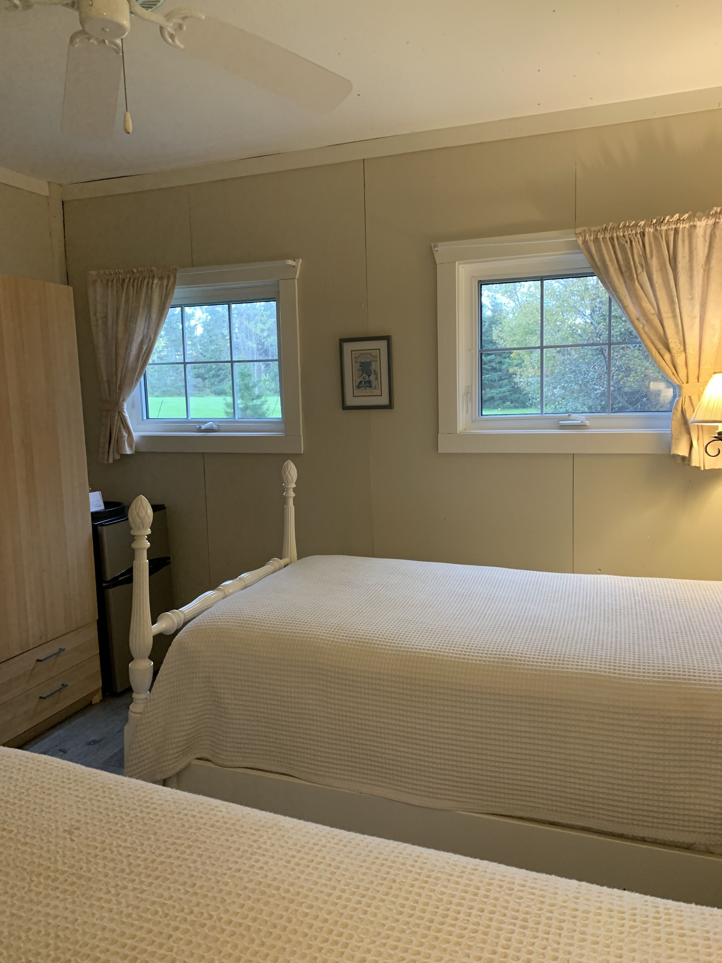 A bedroom with two beds, a ceiling fan, a window with curtains, and a framed picture on the wall.