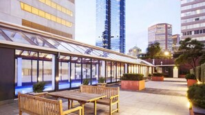 An outdoor seating area with wooden furniture and a glass enclosure, surrounded by modern buildings. - Central 2BR Apt in Southbank (Southbank)