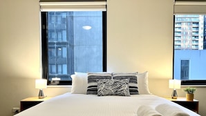 A hotel room with a large bed, two bedside tables with lamps, a zebra-patterned pillow, and a view of buildings through the window. - Central 2BR Apt in Southbank (Southbank)