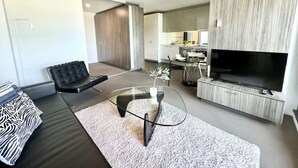 A modern living room with a sofa, a glass coffee table, a flat-screen TV, and a kitchen in the background.