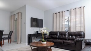 Ein Wohnzimmer mit braunem Ledersofa, einem runden Holztisch mit weißer Vase, einem wandmontierten Flachbildfernseher und einem Fenster mit beige farbenen Vorhängen.