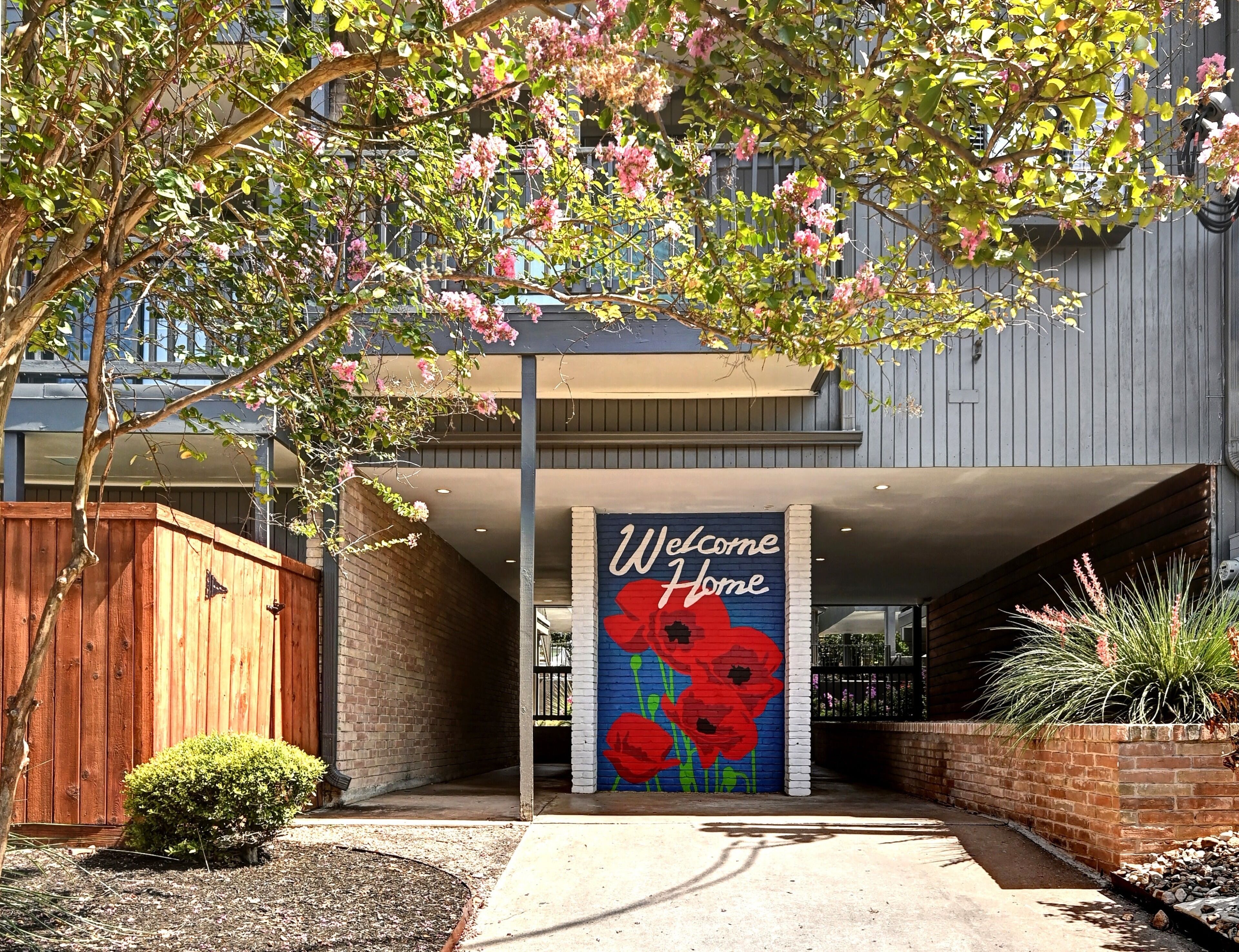 A building entrance with a 'Welcome Home' sign, a wooden fence, and a small bush.