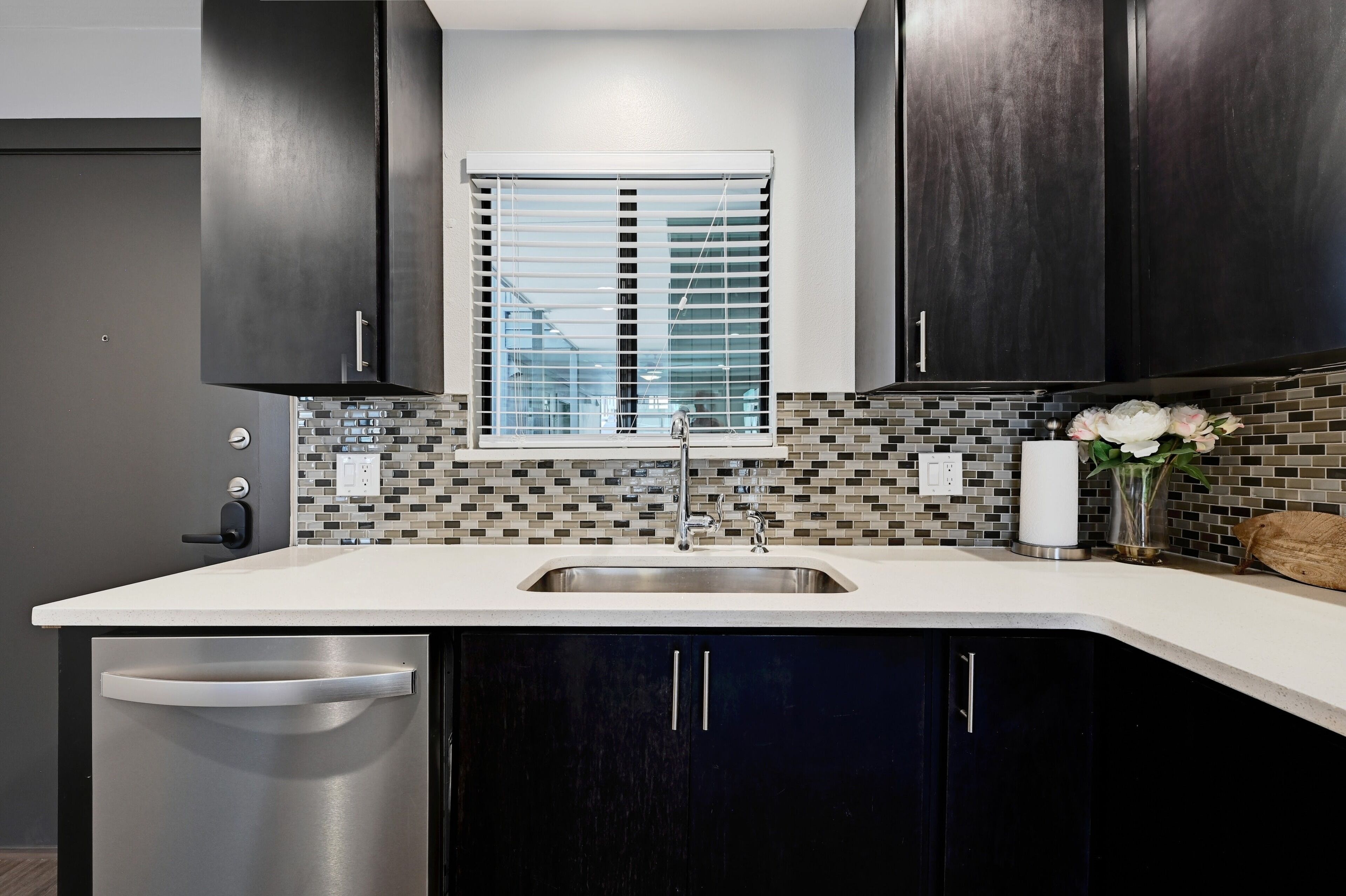 A modern kitchen with dark cabinets, a white countertop, a stainless steel sink, and a window with blinds.